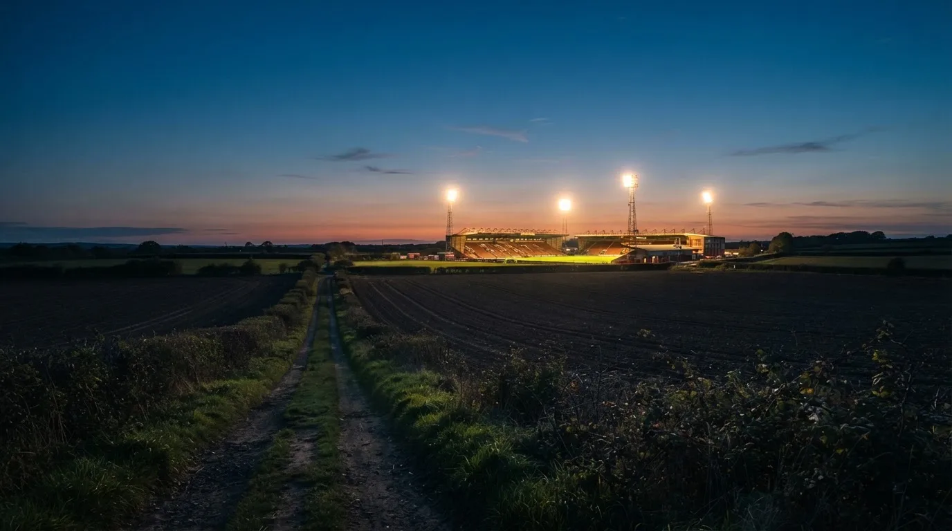 Stadion rozświetlony reflektorami widziany z daleka znad ciemnych pól w pogodny wieczór.