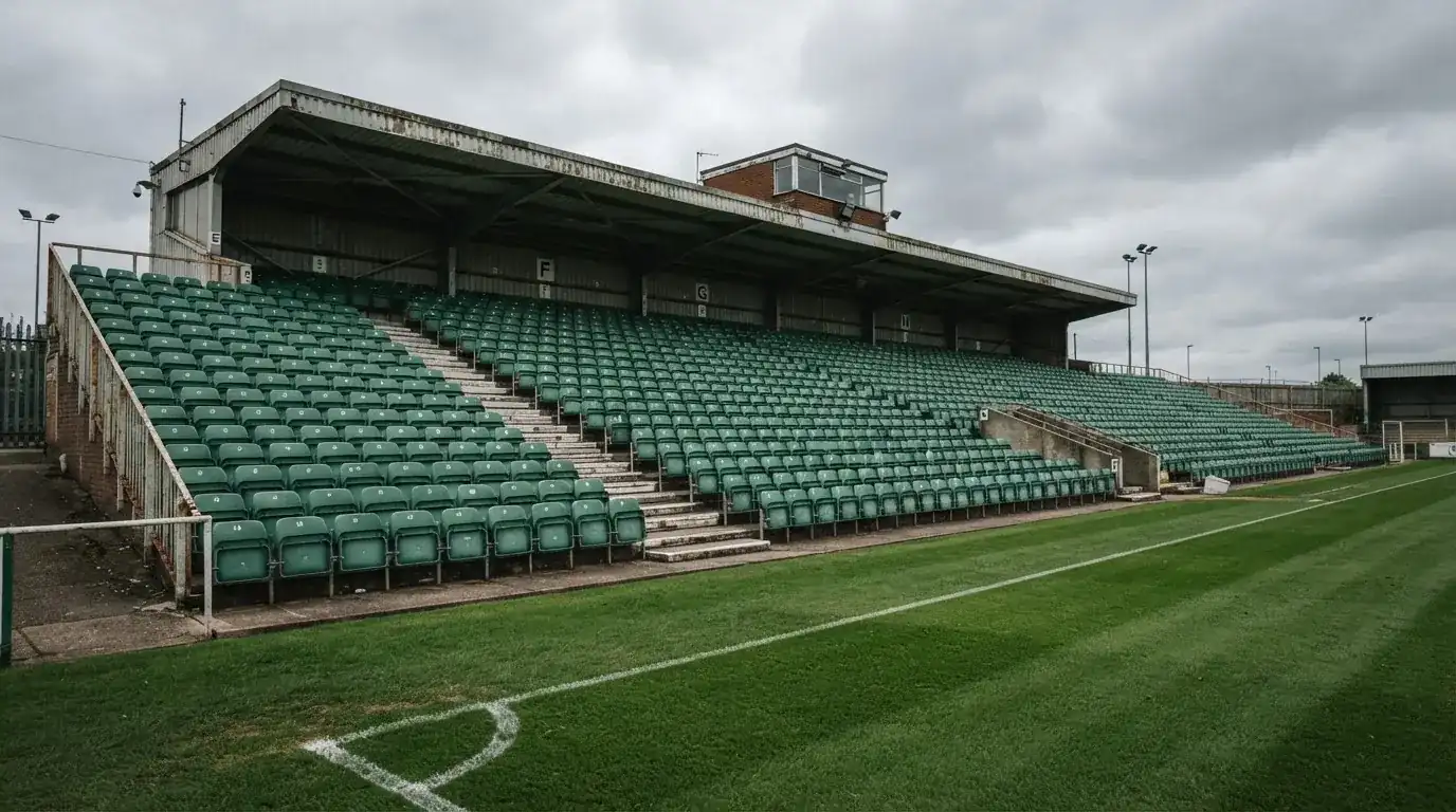 Rzędy pustych plastikowych krzesełek małego stadionu widziane z poziomu murawy pod pochmurnym niebem.