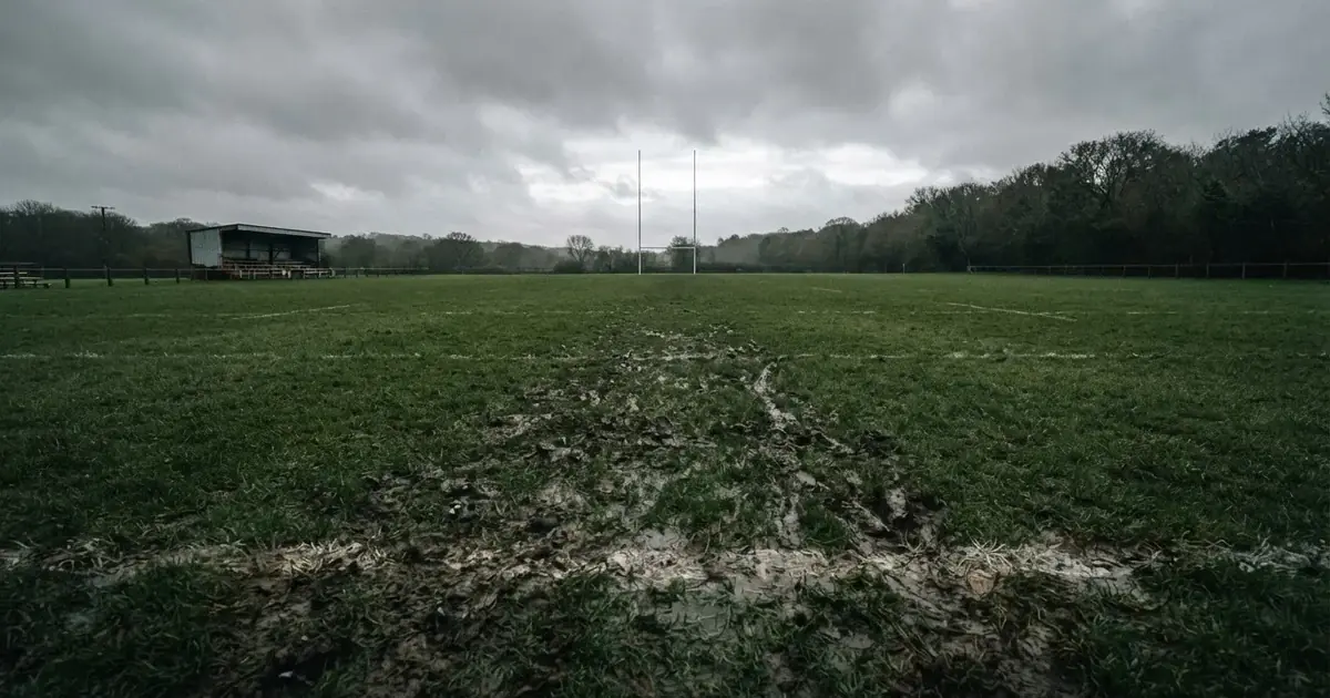 Boisko do rugby ze słupami bramki w oddali pod pochmurnym niebem, mokra trawa i ślady butów.