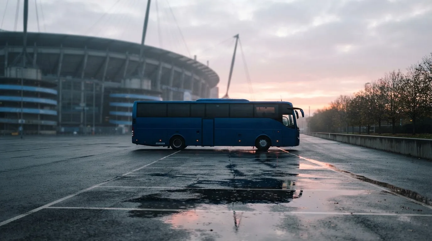 Autokar stoi na parkingu przed stadionem o świcie, rozmyte kontury trybun w tle, pusty plac i mokry asfalt.